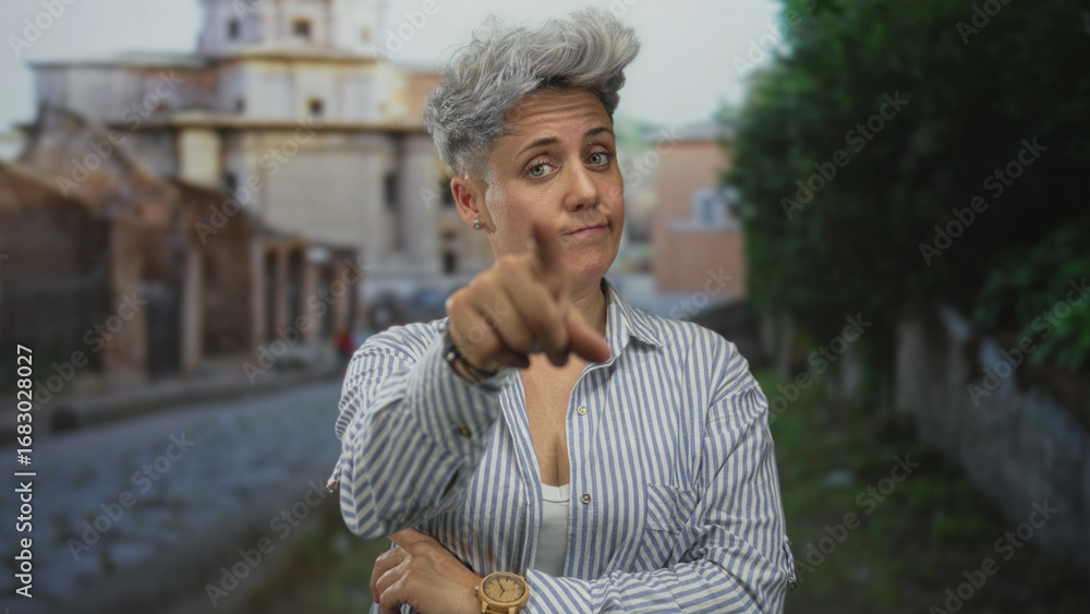 Fototapeta premium Woman with gray hair in striped shirt finger pointing forward on street; doubt uncertainty reflection.