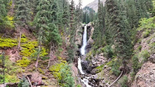 Drone approaches Barskoon waterfall in Kyrgyzstan, then ascends to reveal a stunning top-down view of the falls, surrounded by forest and mist in the mountains.