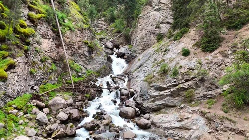 A slow drone flight starts over a stream running through rocks and gradually rises, revealing the Barskoon Waterfall hidden among the trees in the mountains of Kyrgyzstan.