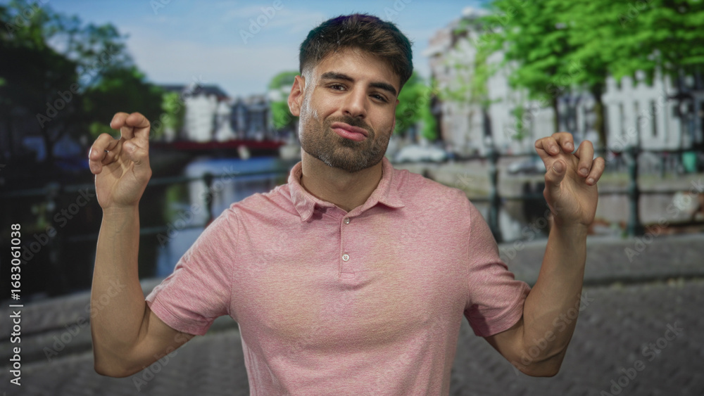 Naklejka premium Young hispanic man in pink polo shirt showing playful smirk with hands making quote gesture on a street; sarcasm.