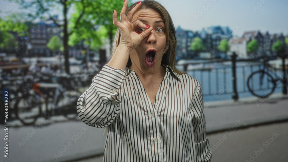 Fototapeta premium Woman gestures ok sign with hand along sunlit canal embankment on a lively bustling city street; approval.