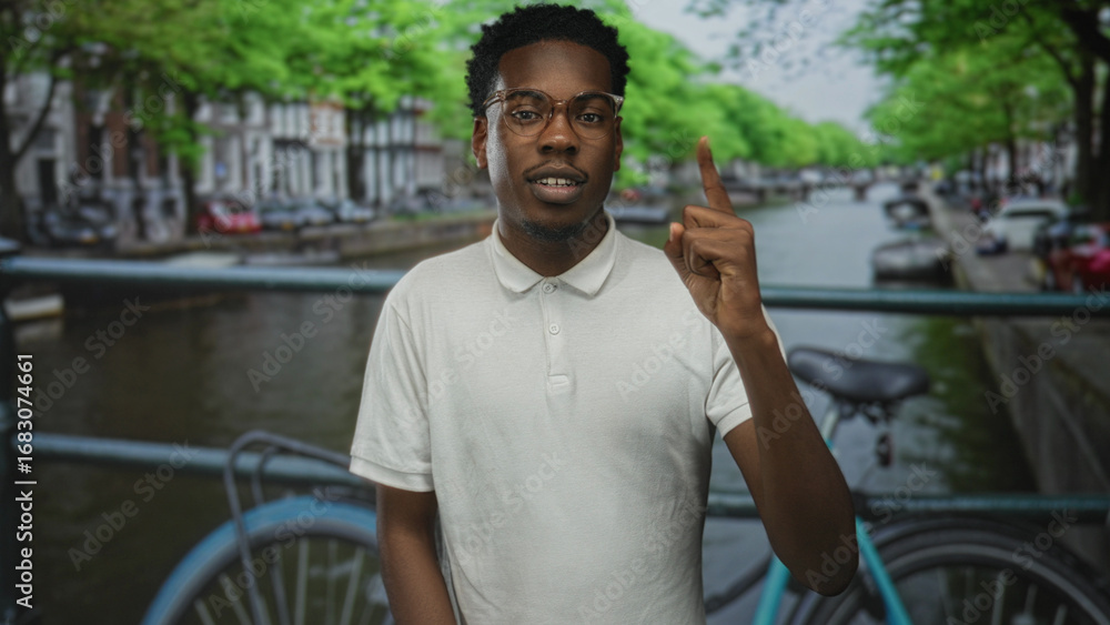 Fototapeta premium Man points finger upward at canal edge in city street framed by leafy trees; curiosity and discovery.