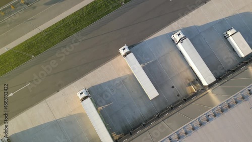 Aerial view of a logistics hub with four white trucks, ready for cargo distribution. The expansive warehouse features solar panels, emphasizing sustainable operations.