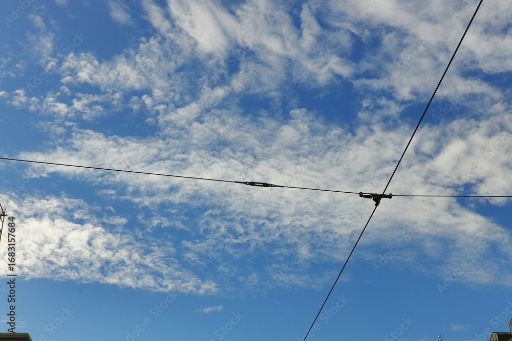 Fototapeta premium Blue sky with white clouds intersected by tram or trolleybus wires, with city buildings below. Urban infrastructure and abstract cityscape concept.