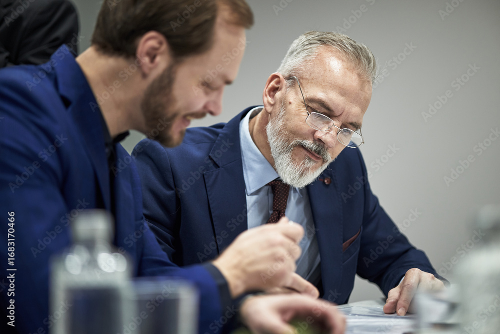 Fototapeta premium Middle aged Caucasian man with gray beard sitting beside young male partner, both wearing suits, reviewing documents together at table in office, conducting negotiations on contract terms