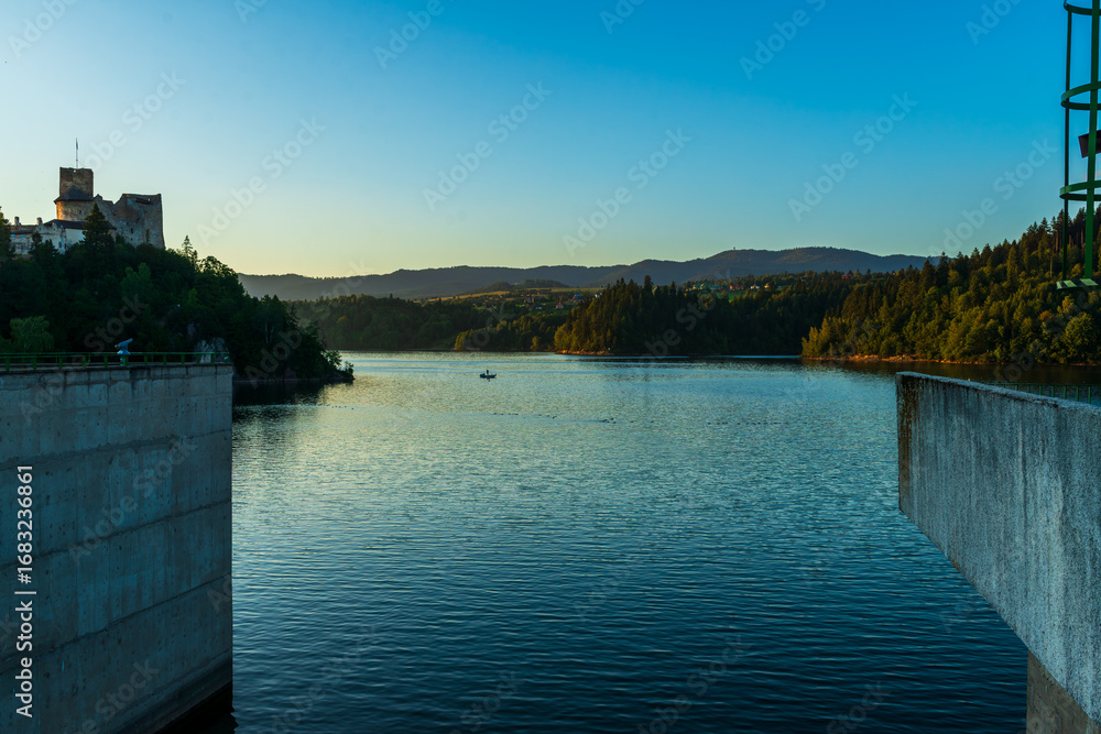 Obraz premium A view of Czorsztyn Lake with Niedzica Castle and distant hills under soft evening light. A boat drifts quietly across the water, emphasizing peace, tranquility, and the harmony of history with nature