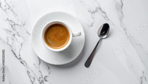 A freshly brewed espresso coffee in a small white ceramic cup and saucer, accompanied by a silver spoon, all elegantly presented on a veined white marble surface.