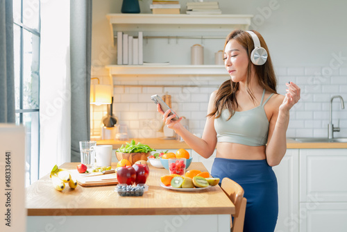 Healthy Asian woman in activewear prepares a fresh fruit drink, slicing fruit and blending a protein shake in her modern pantry kitchen at home, promoting wellness and fit lifestyle	