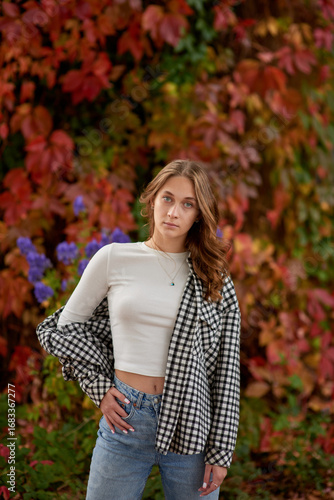 Young woman in casual autumn outfit posing against vibrant red ivy wall, stylish fashion portrait in warm seasonal tones.