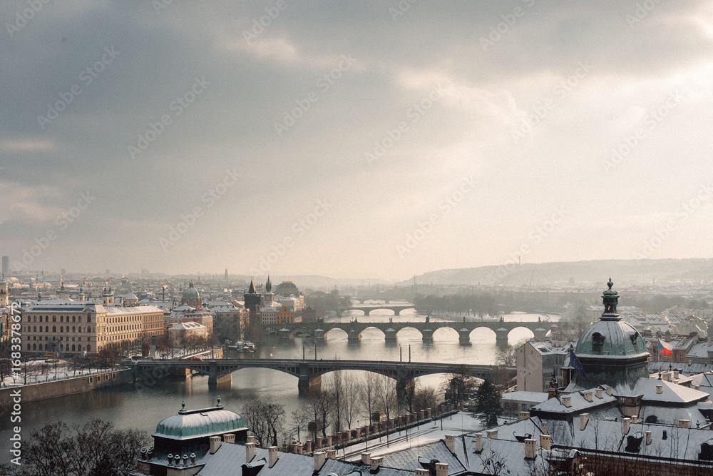 Fototapeta premium A stunning winter scene of Prague, showcasing snow-covered rooftops and bridges over the Vltava River, illuminated by soft sunlight.