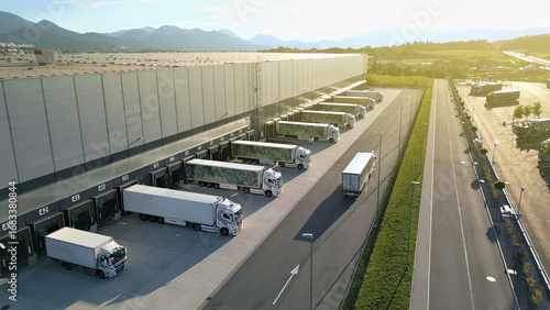 Aerial view of a busy distribution warehouse with trucks at loading docks, set against scenic mountains. Highlights efficient freight transportation and logistics, modern infrastructure.
