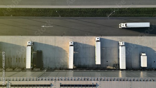 Aerial view of a distribution warehouse with five cargo trucks at loading docks and one on the road, showcasing efficient freight logistics and organized cargo handling.