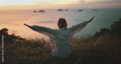Woman sit grassy hilltop with arms outstretched, meet sunrise over ocean. She enjoy peaceful scenery and warm glow of morning light. Freedom and tranquility, beauty of Thailand tropical island nature