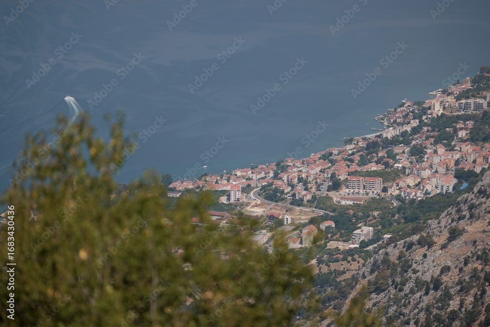 Obraz premium A high angle shot of a coastal European town with terracotta rooftops nestled between mountain and sea. A speedboat creates a bright wake on the dark water of the Bay of Kotor