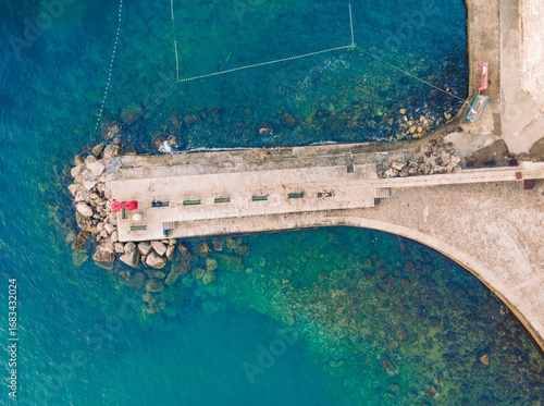 Captured from above, benches line a stone walkway near a harbor in Split. Azure waters embrace the shoreline, and sunlight dances on the water surface, revealing rocks underneath