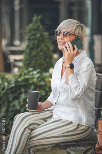 40s middle age blond woman on the mobile phone talking and resting on bench in the city. Side view of adult female talks on the smartphone.