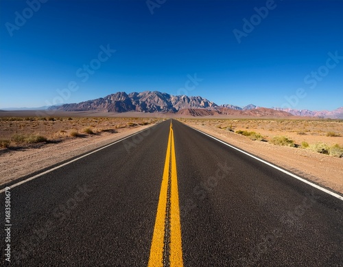 an empty straight road stretches towards a majestic mountain range under a clear blue sky the golden hues of the desert contrast with the striking double yellow lines of the road inviting viewers