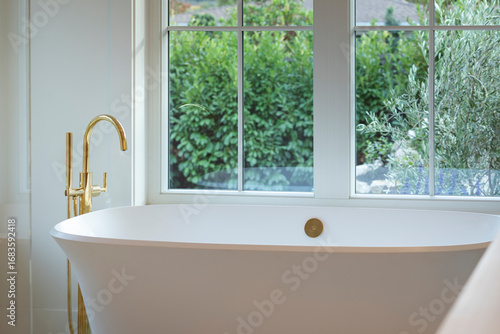 Relaxing scene of soaking tub with gold faucet and view of greenery.