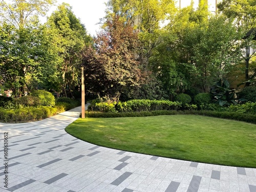 Cuadro en lienzo Landscaped residential or commercial complex with paved walkway, manicured lawn, trimmed shrubs, tall green and red-leaved trees, and woven outdoor seating in a clean, comfortable setting