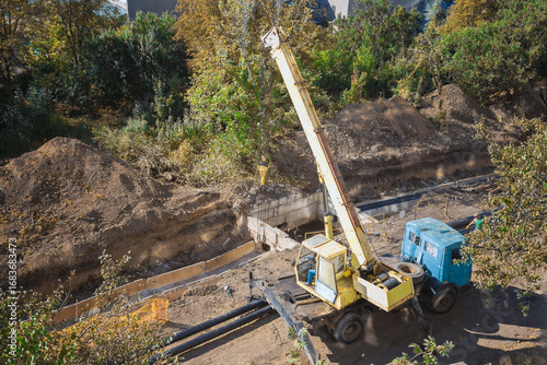 Mobile crane on sunlit construction site among trees, construction and repair works using heavy machinery, break in work at construction site.