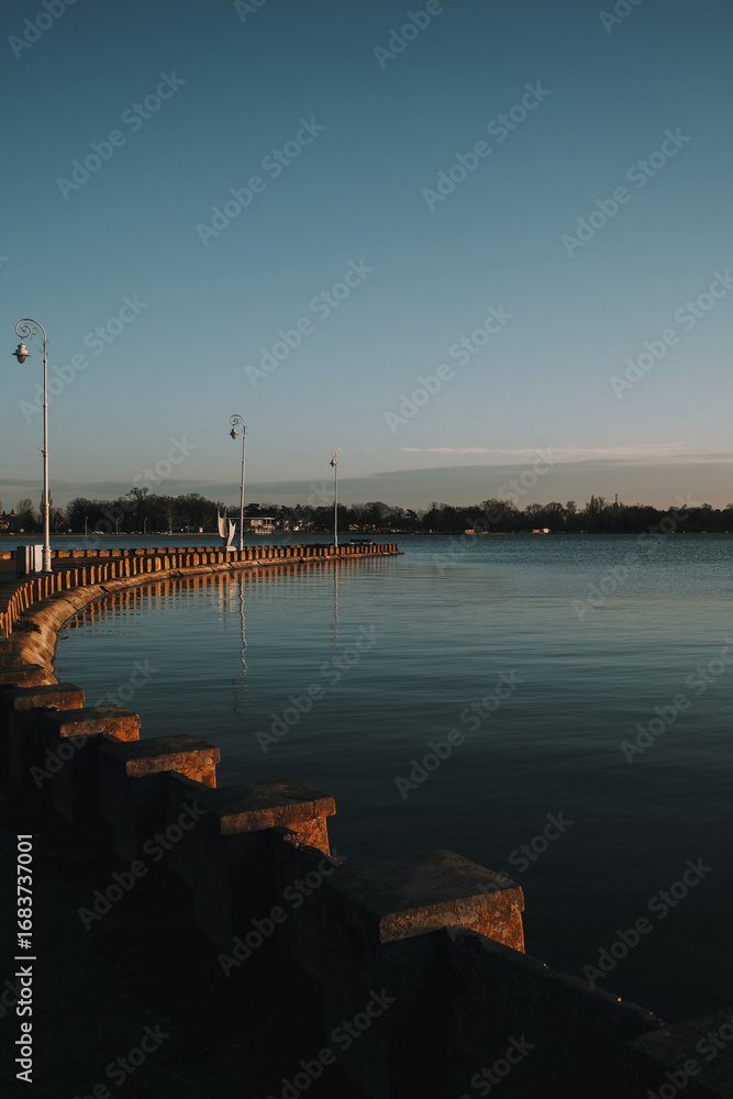 Obraz premium A quiet winter evening at Palic Lake promenade in Serbia, with vintage street lamps casting long shadows over the calm water