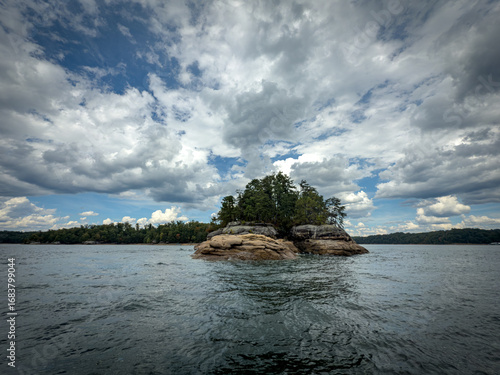 Rocky sandstone outcrop with dense trees surrounded by the waters of Laurel River Lake, Kentucky. A peaceful wilderness scene with textured rock formations, lush forest, and striking cloudy skies.