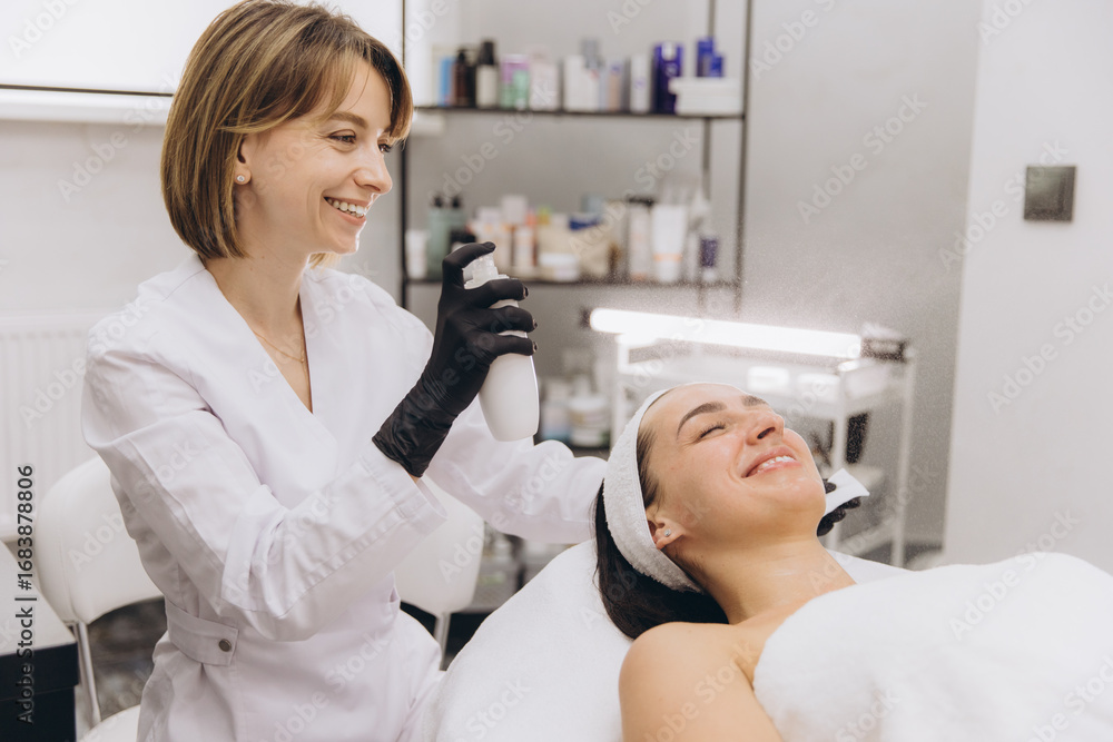 Fototapeta premium Beautician spraying moisturizing treatment on face of woman lying on bed
