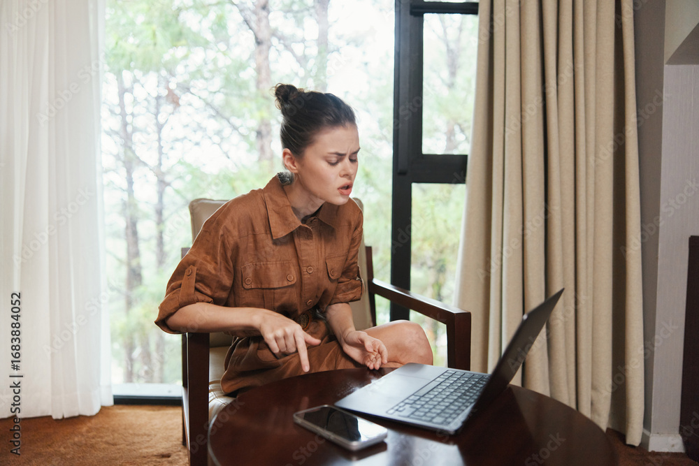 Fototapeta premium Young woman in brown dress sitting by a window, expressing stress while working on laptop, modern home office setting with natural light