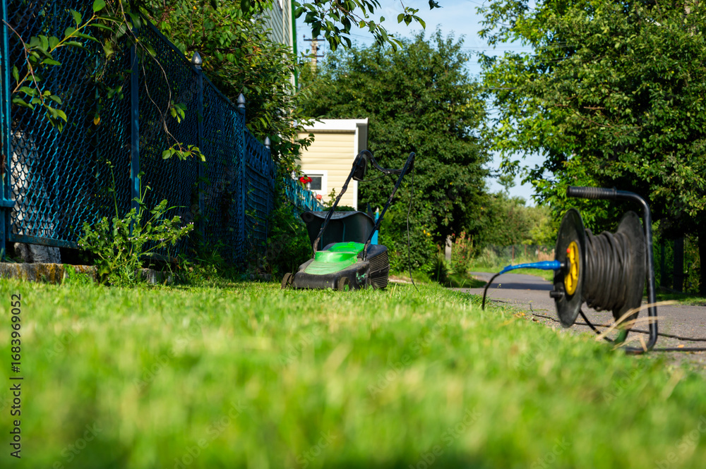 Fototapeta premium Mower sits on freshly cut grass in a residential setting under clear blue skies, with green trees lining the walkway