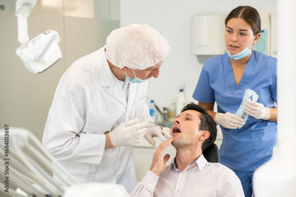 Obraz premium Man sits in armchair during visit to senior male dentist and undergoes diagnostic procedure. Doctor examines patient and asks for help of girl nurse, gives instructions on how to prepare instrument