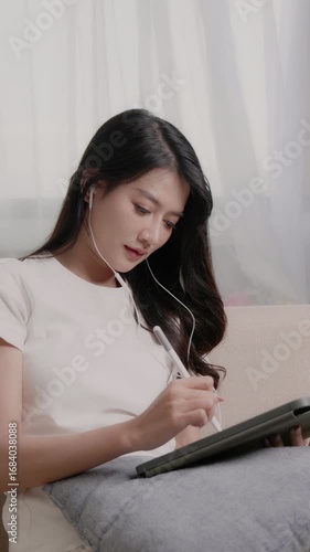 woman in casual clothes is writing something on a tablet while sitting on a sofa in a living room.