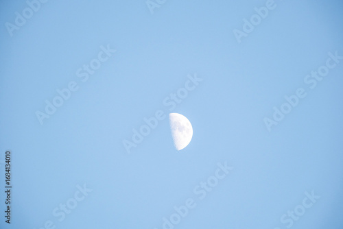 Fototapeta Naklejka Na Ścianę i Meble -  Nasa The first quarter half moon hangs clearly visible in a vast, bright, and minimalist pale blue daytime sky. The serene, simple composition emphasizes solitude and celestial presence in the dayligh