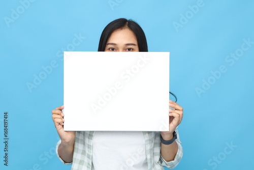 Young Asian woman wearing casual clothes, holding blank white paper board for use as advertisement, standing isolated on blue background.