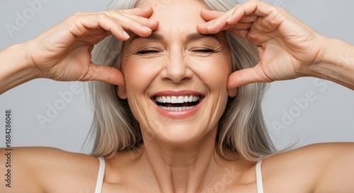 An ecstatic senior woman with gray hair laughs while stretching her facial skin