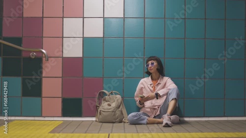 A young woman is sitting at a subway station and waiting for a train. A passenger on the platform is waiting for a train and using a phone. The girl is looking at the schedule.