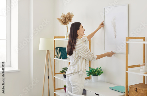Fotografie Cheerful young woman arranging piece of art on wall, hanging picture on wall in her sunny, contemporary apartment, smiling and organizing, decorating her living space