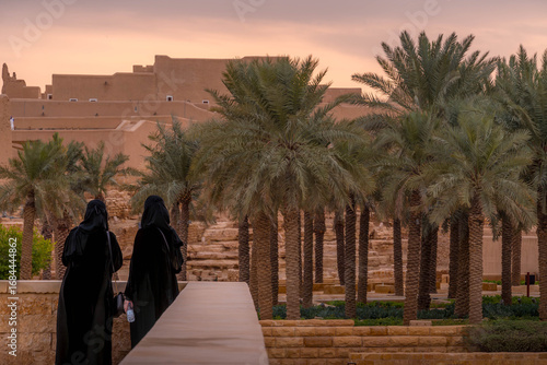 Two Saudi women in Diriyah courtyard, wearing traditional abayas and hijabs, among restored Najdi mudbrick architecture.
