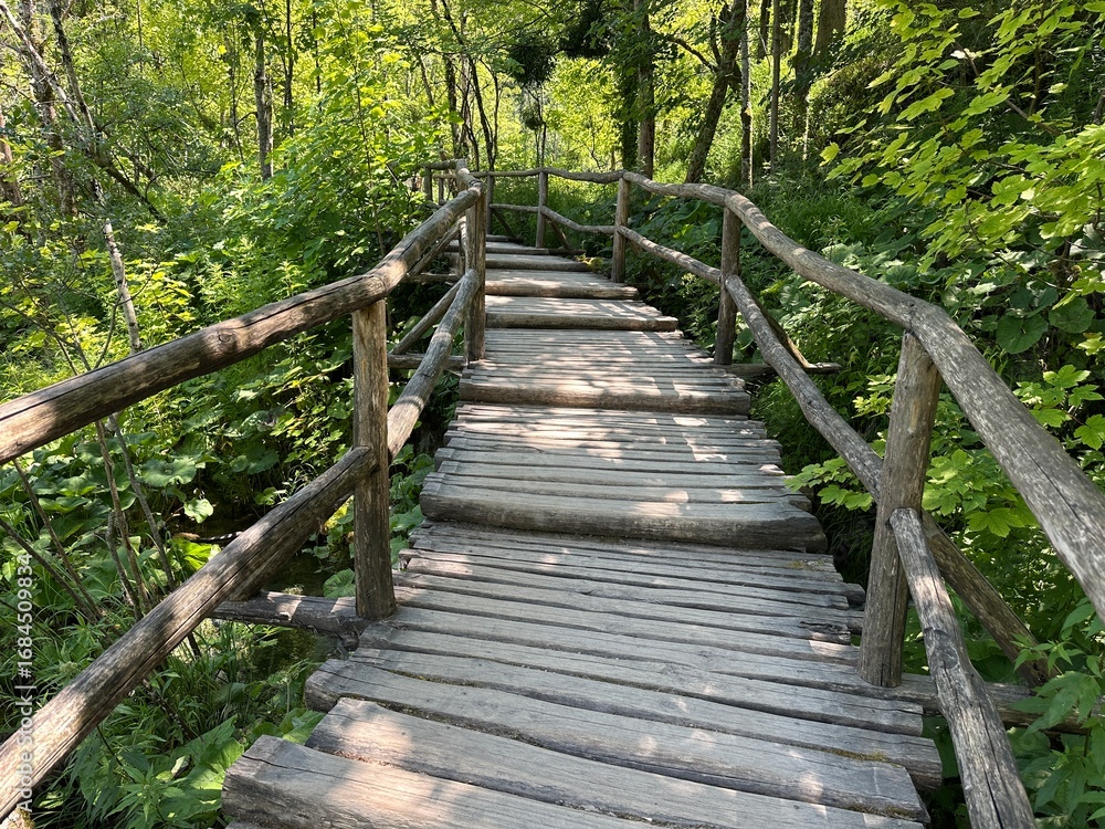 Obraz premium Experience the beauty of Plitvice Lakes with its iconic wooden bridge, surrounded by sparkling blue waters and magnificent waterfalls—a perfect serene escape into nature's embrace.