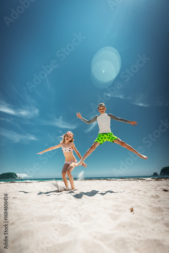 Children leap into summer sky on sunny beach, full body jump frozen mid air. Pure childhood freedom and energy against vivid blue backdrop. Early travel booking