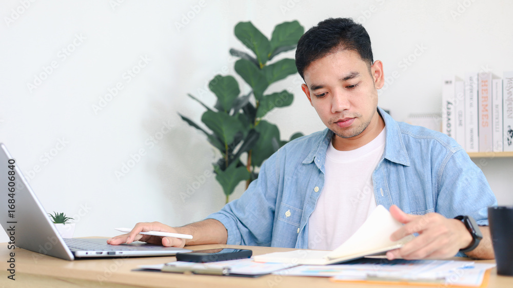 Fototapeta premium Young Asian man working on laptop at home office desk with books and plants in background. Concept of remote work, freelance business, and digital lifestyle.