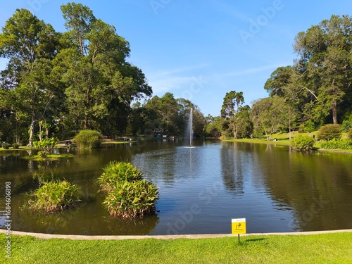 A serene lake surrounded by lush greenery, trees, and a fountain under a clear blue sky.