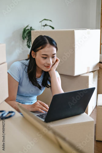 Female Asian use on laptop surrounded by moving boxes at home