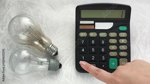 An incandescent lamp is lying on a table, a woman is counting the price of electricity on a calculator. The concept of rising electricity prices, economics