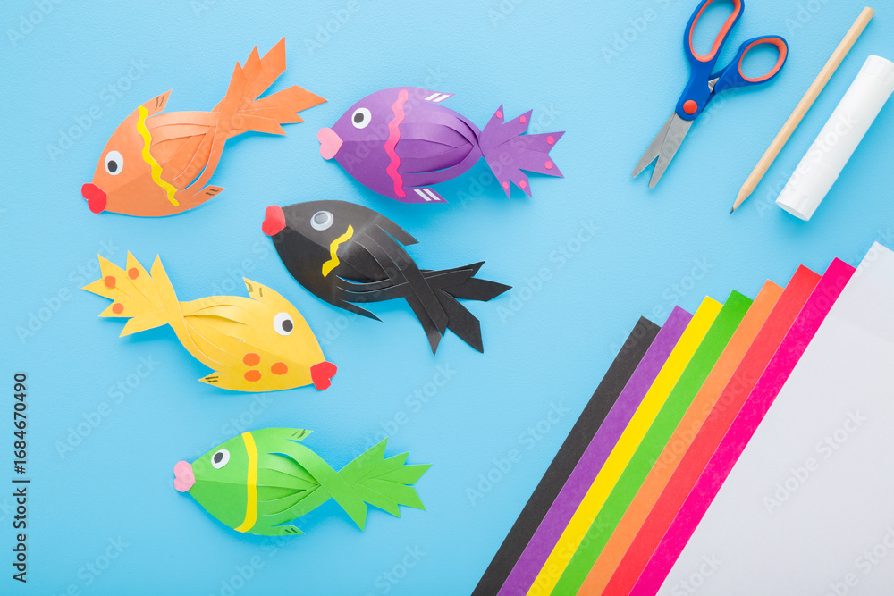 Fototapeta premium Fishes, scissors, glue stick, pencil and colorful application paper on light blue table background. Pastel color. Closeup. Child created shapes. Top down view.