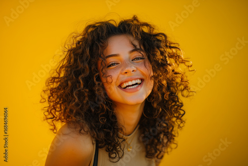 Joyful young woman with curly hair laughing against vibrant yellow background in natural light