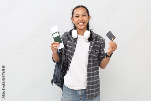A young Asian male student wearing casual clothes, backpack, headphones around his neck, smiling facing the camera while holding passport ticket and credit card, school holiday concept.