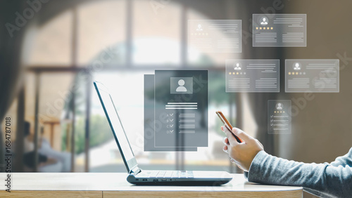 A person uses a smartphone to review digital candidate profiles and ratings on a holographic interface above a laptop in an office.