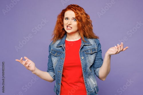 Young sad ginger woman wears red t-shirt denim shirt shrugging shoulders looking puzzled, have no idea look camera isolated on plain pastel light purple background studio portrait. Lifestyle concept.