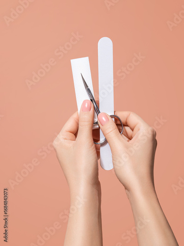 Hands with nail buffer block and manicure scissors on peach background
