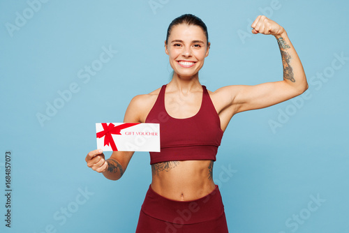 Young fitness trainer instructor sporty woman in red sportswear train in home gym hold gift coupon voucher card for store show muscles isolated on plain blue background Workout sport fit abs concept